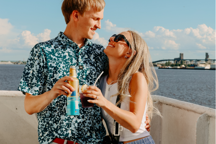 Two people smiling happily on a boat holding drinks, with water and a bridge in the background.