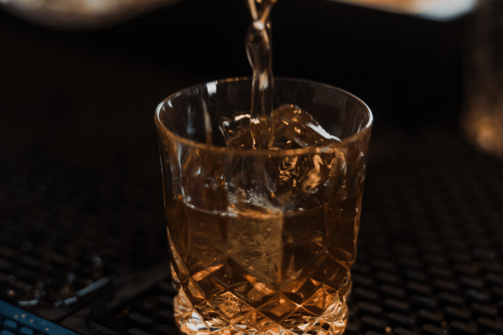 Amber liquid being poured into a glass with ice cubes on a bar mat.