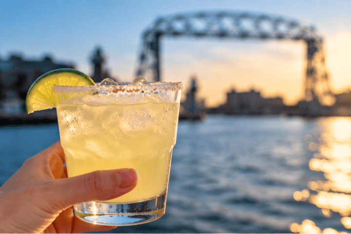 Hand holding a lime cocktail with a bridge and sunset in the background.